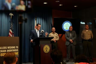 Hestrin, wearing a blue tie and glasses, speaks into a microphone on a podium with the Riverside DA's seal.