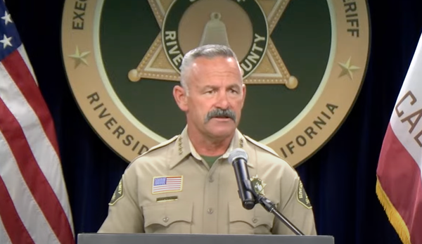 Sheriff Bianco, in tan uniform, speaks in front of a microphone, backed by his sheriff's seal, and flanked by the California and American flags
