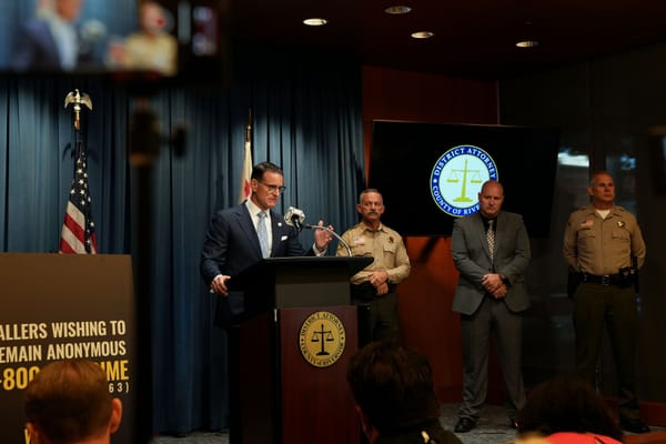 Hestrin, wearing a blue tie and glasses, speaks into a microphone on a podium with the Riverside DA's seal.